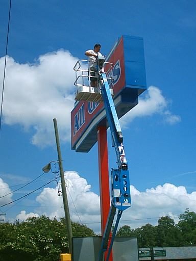 Leo on a lift is repairing a tall sign against a clear blue sky.